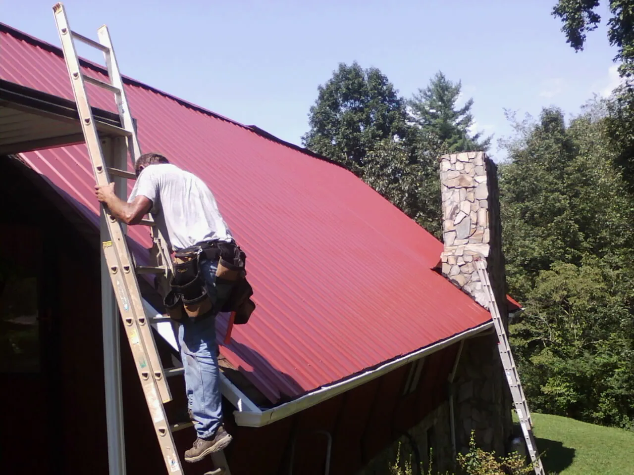Roofer on a ladder beside a red metal roof and stone chimney — Soffit & Fascia Repair by Roofing Brothers in West Harrison